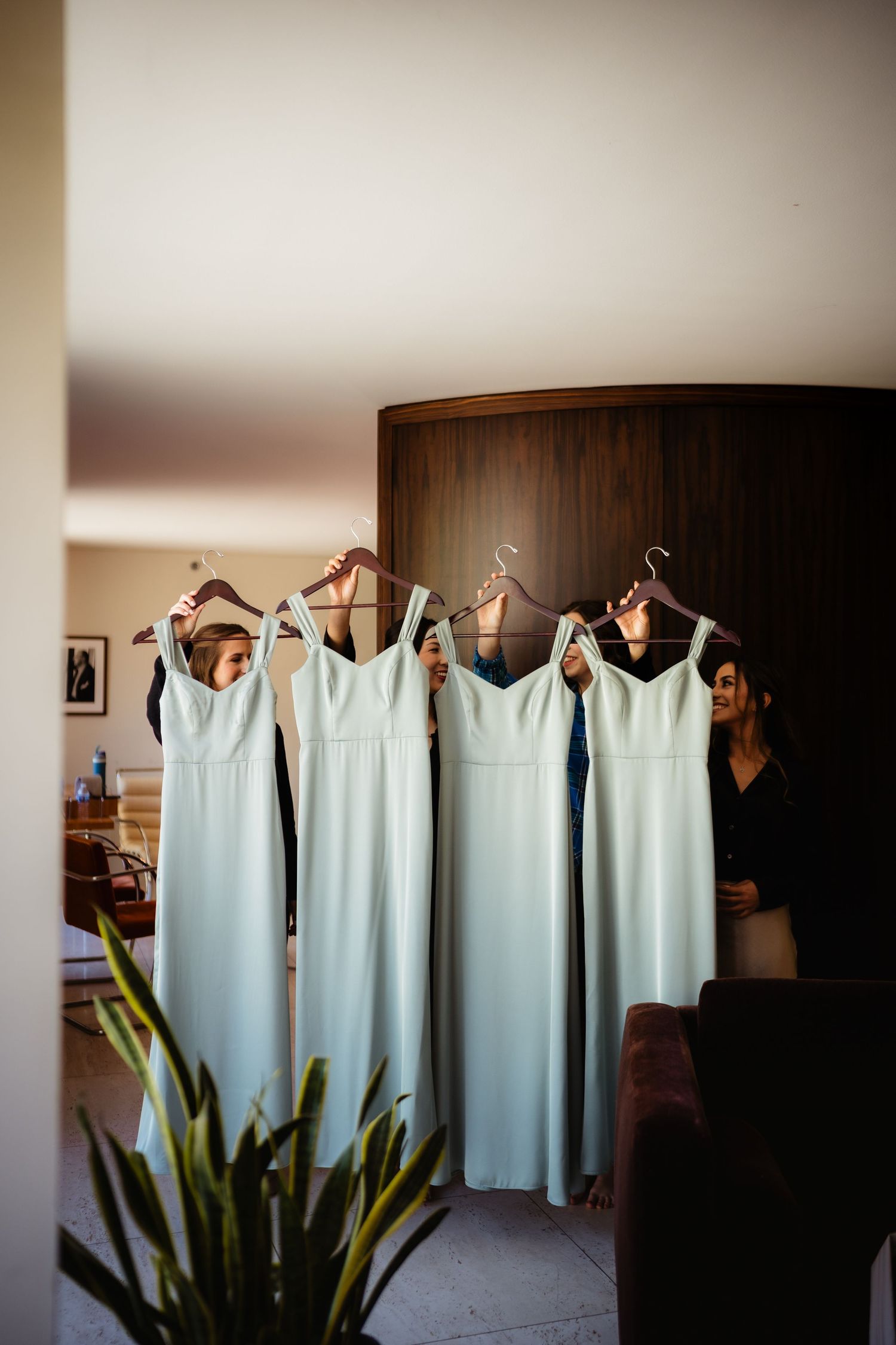 Bridesmaids holding up four matching sage green dresses on hangers in a modern hotel room before a wedding.