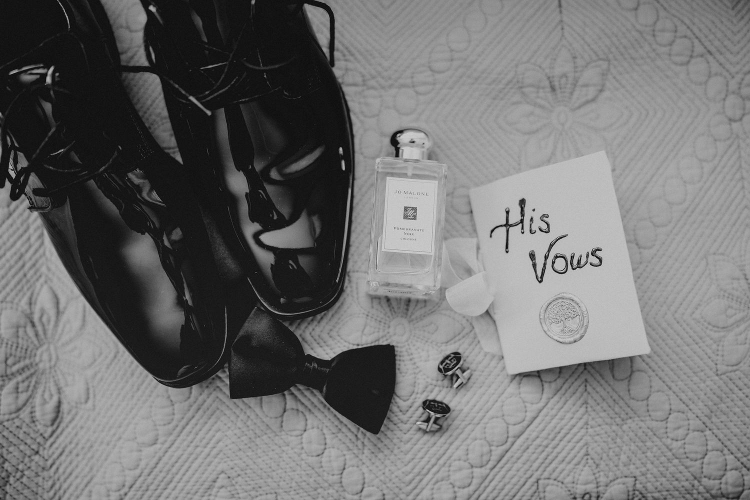 Black and white flat lay of groom wedding essentials including dress shoes, bow tie, cufflinks, cologne, and His Vows booklet.
