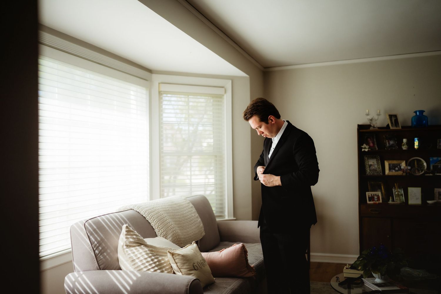 Groom in black suit adjusting his tie in a softly lit living room before the wedding ceremony.