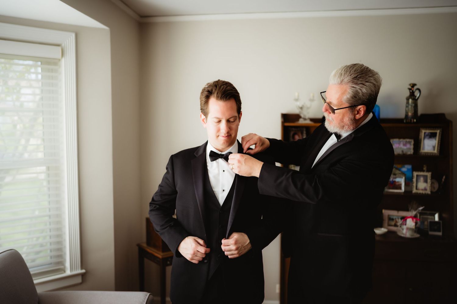 Groom in black tuxedo getting bow tie adjusted by older man in matching tuxedo before wedding ceremony.