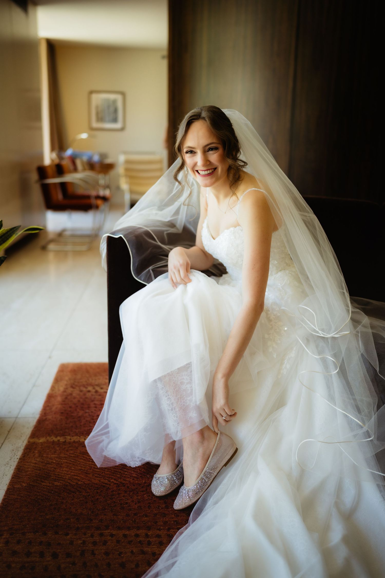 Smiling bride in white gown and veil sits on sofa adjusting her silver shoes before her wedding ceremony.