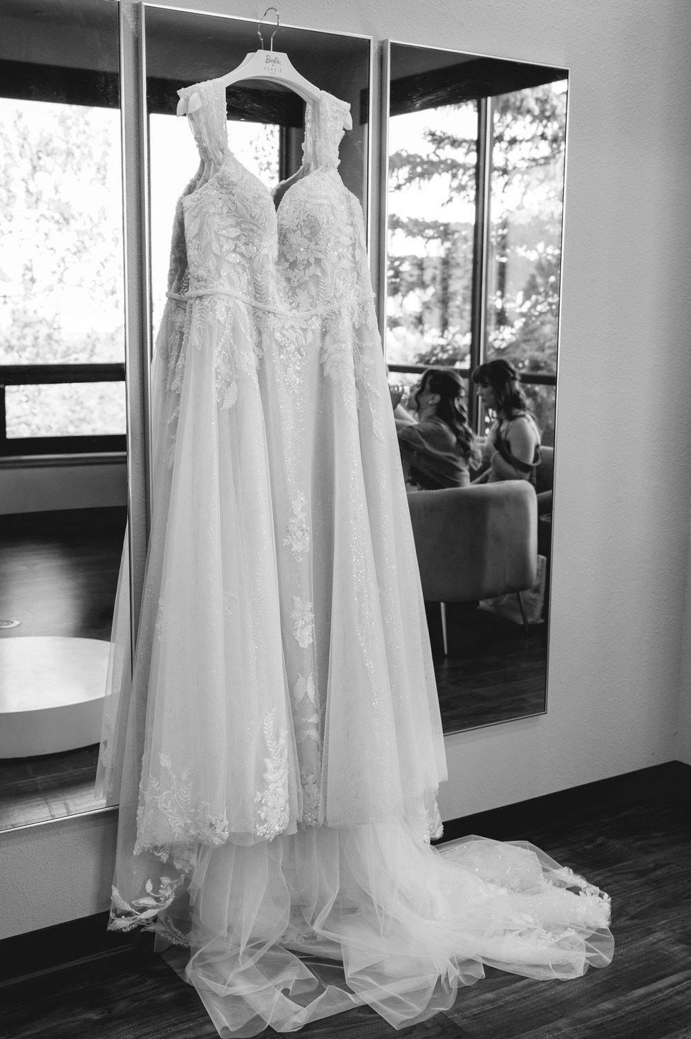 Elegant lace wedding dress hanging on a mirror in black and white, with bridesmaids reflected in background.