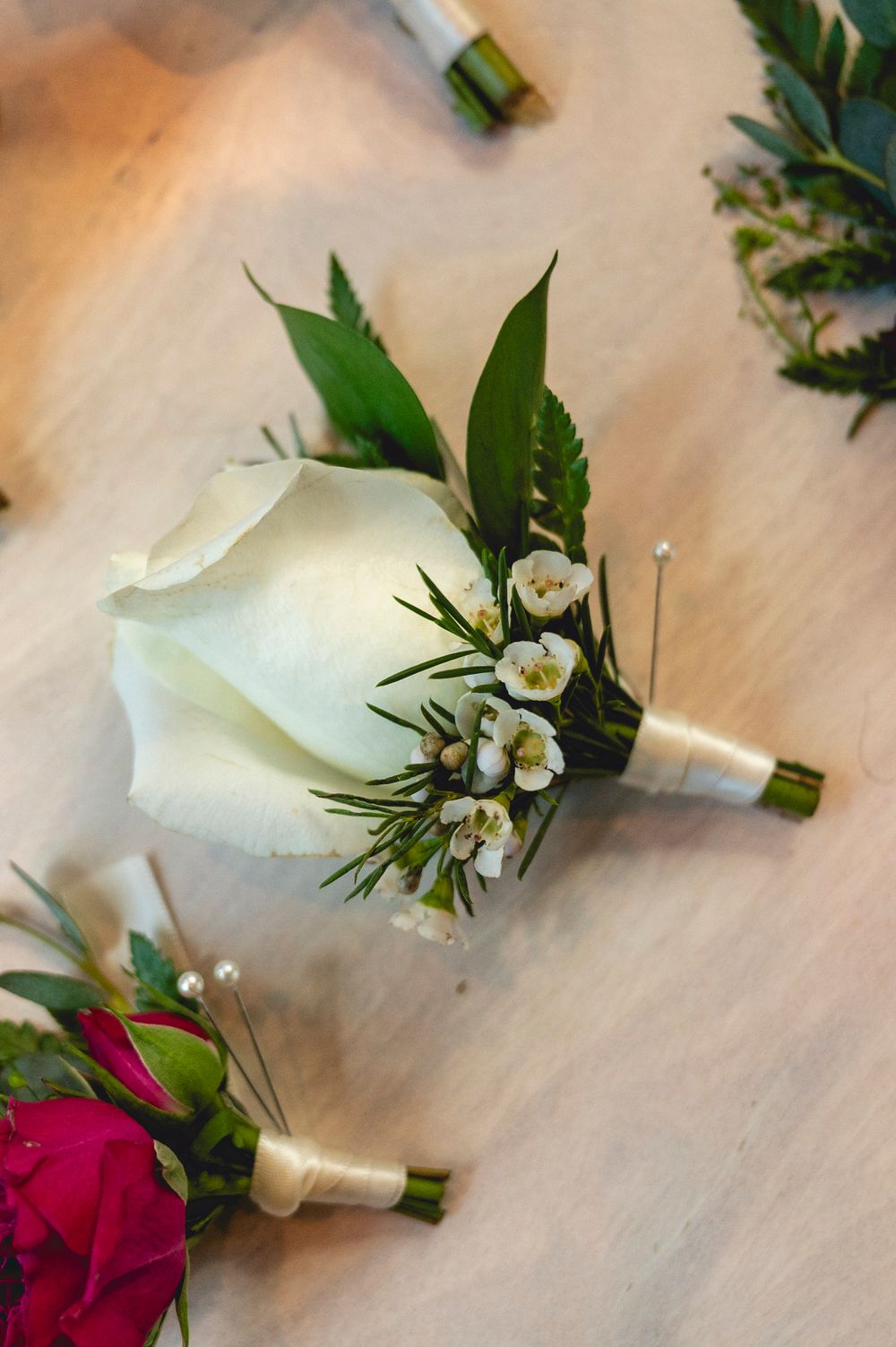 White calla lily boutonniere with wax flowers and greenery beside a pink rose boutonniere on a wooden surface.