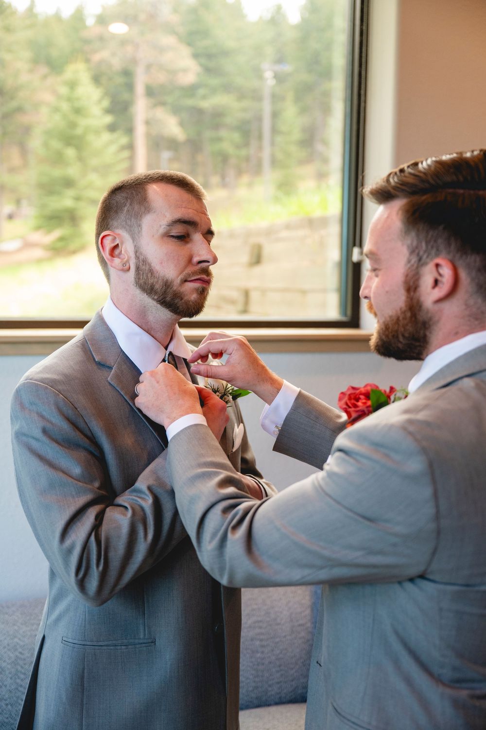 Two groomsmen in gray suits, one pinning a red boutonniere on the other before a wedding ceremony.