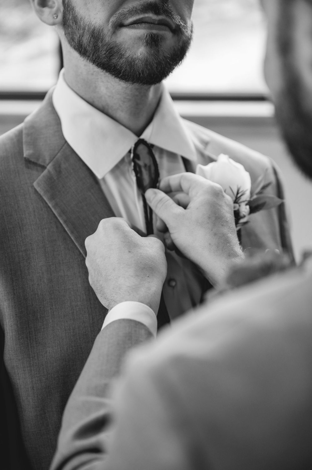 Groom in gray suit gets boutonniere pinned and tie adjusted by groomsman before wedding ceremony.