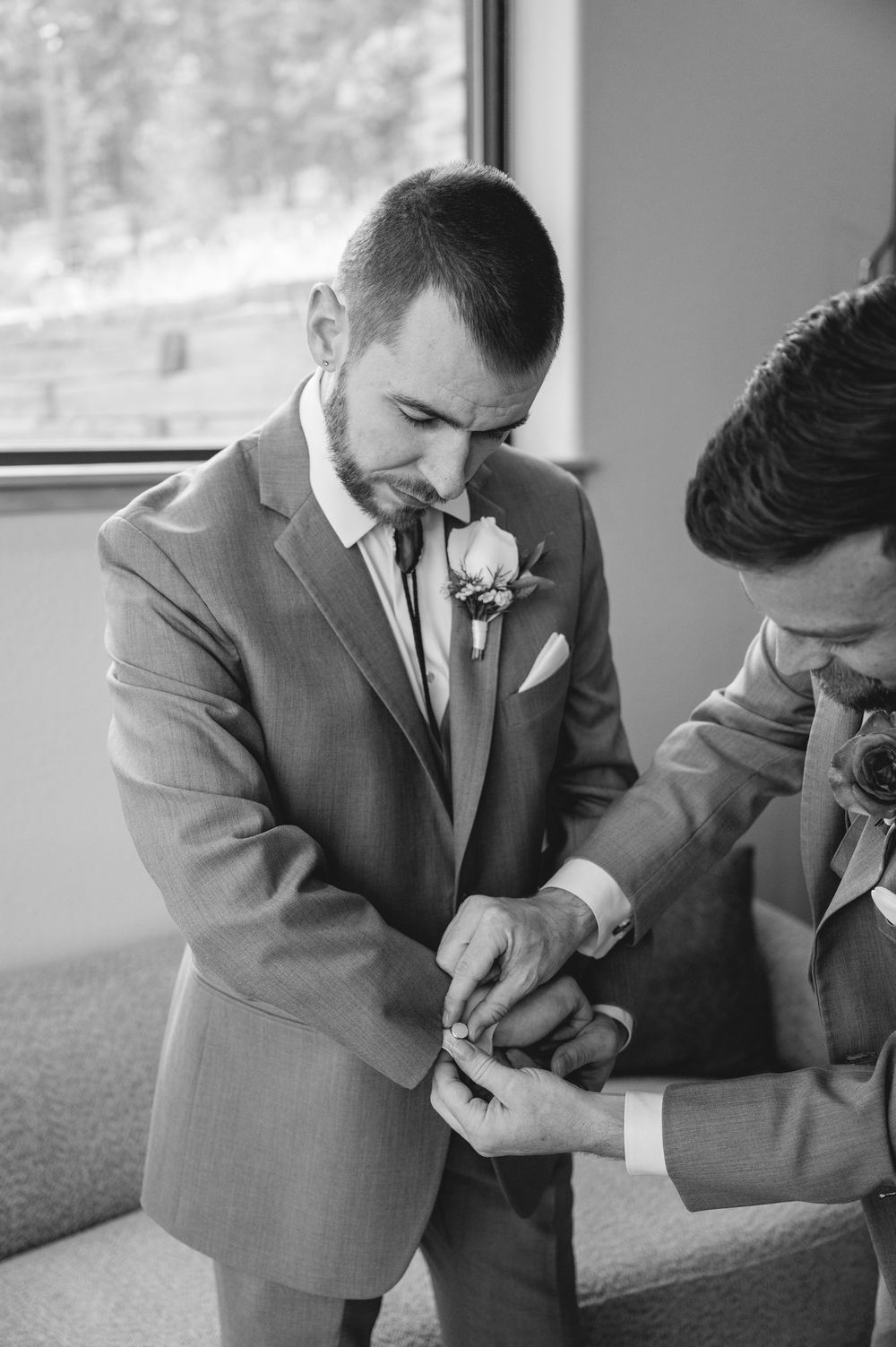 Groom in gray suit with rose boutonniere gets cufflinks fastened by groomsman before wedding ceremony.