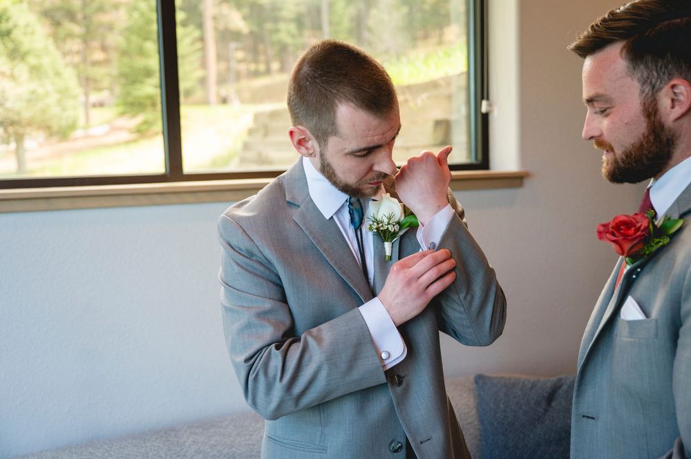 Two groomsmen in gray suits with boutonnieres adjusting cufflinks before wedding ceremony indoors.