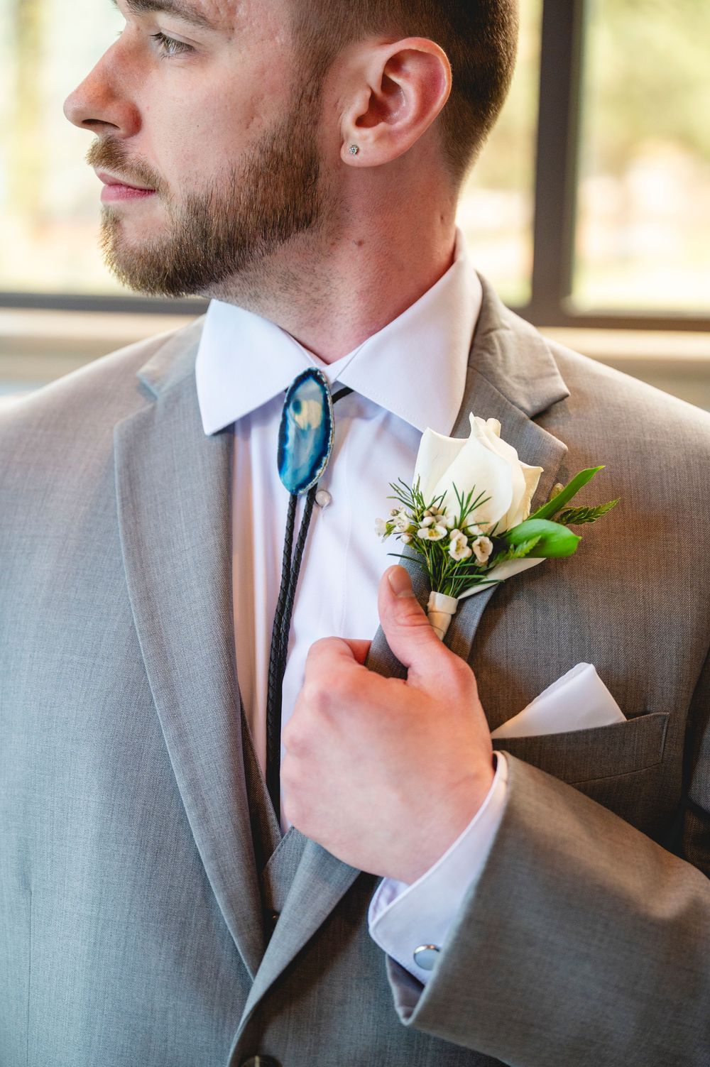 Groom in gray suit adjusts white rose boutonniere wearing unique blue geode bolo tie before wedding.