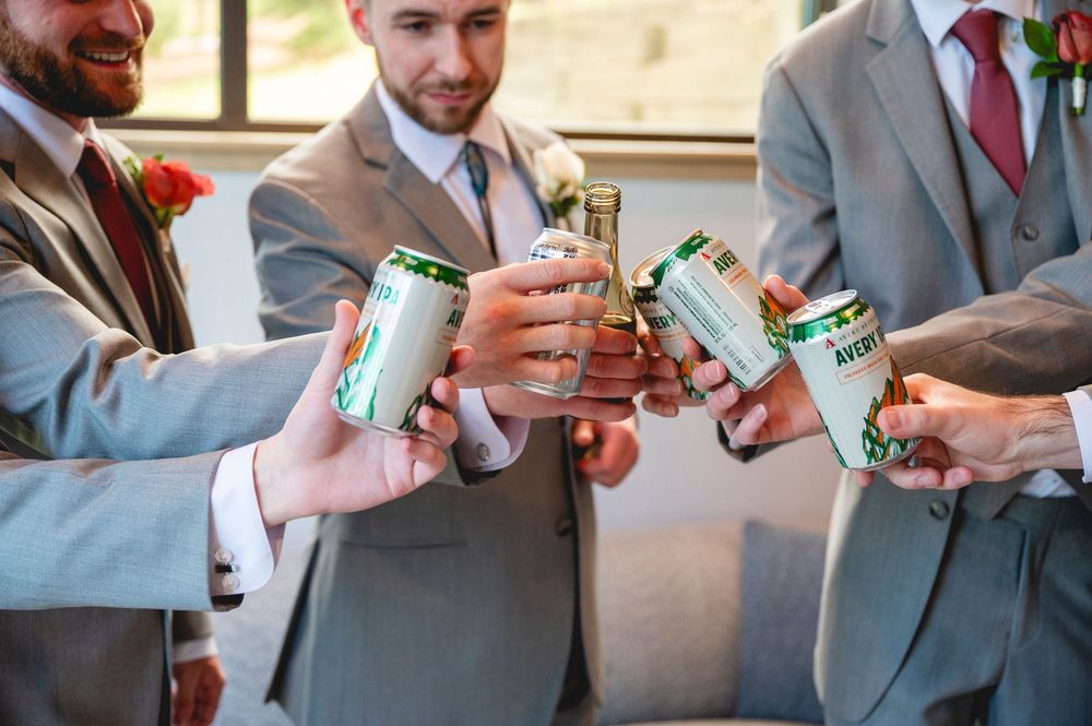 Groomsmen in gray suits toasting with beer cans at a wedding celebration.