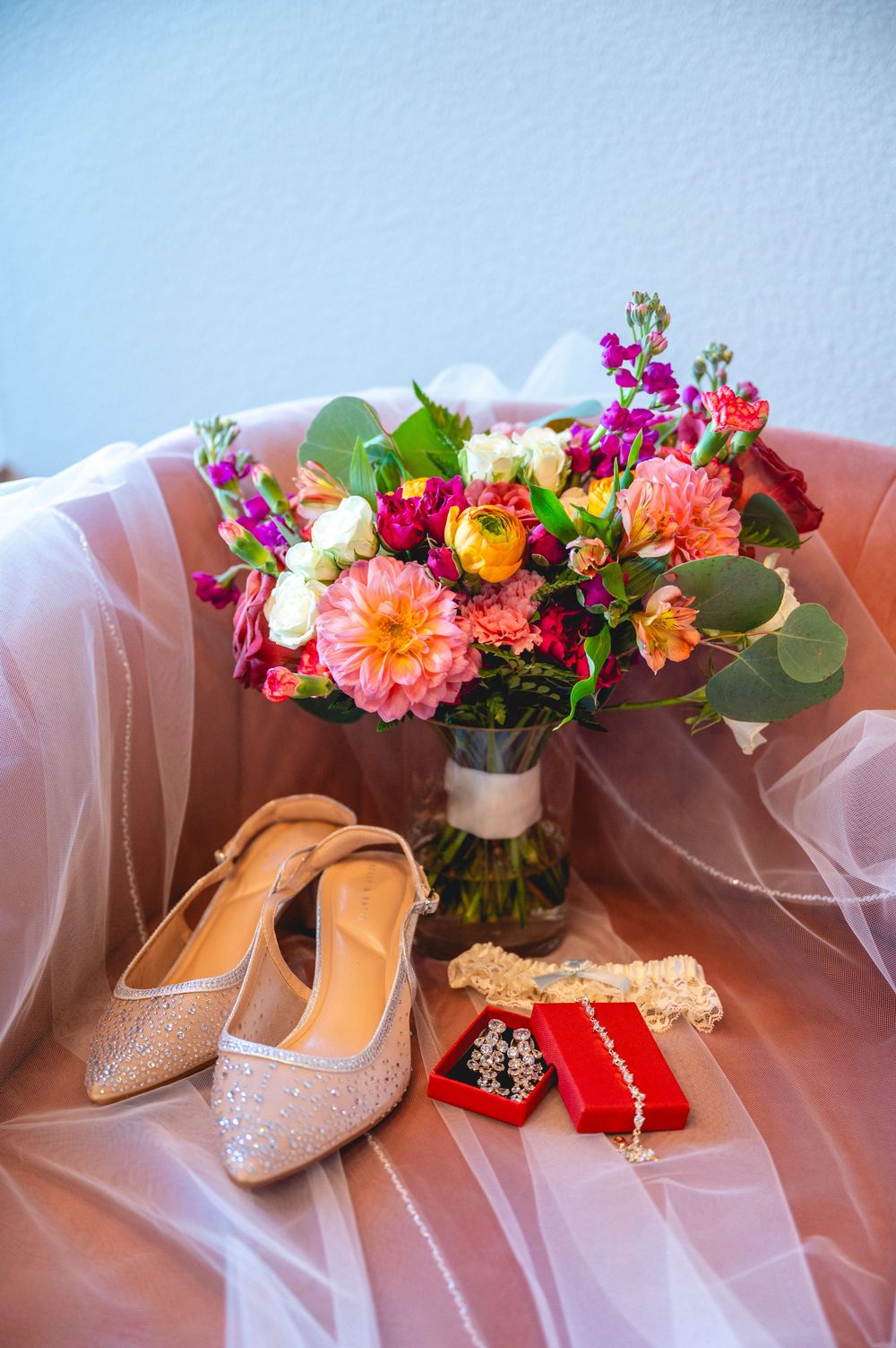 Bridal flat shoes, colorful bouquet with dahlias, jewelry in red box, and garter on pink tulle fabric.