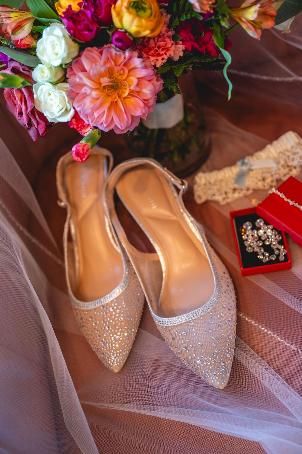 Elegant gold rhinestone slingback flats beside a vibrant bridal bouquet and jewelry on soft tulle fabric.