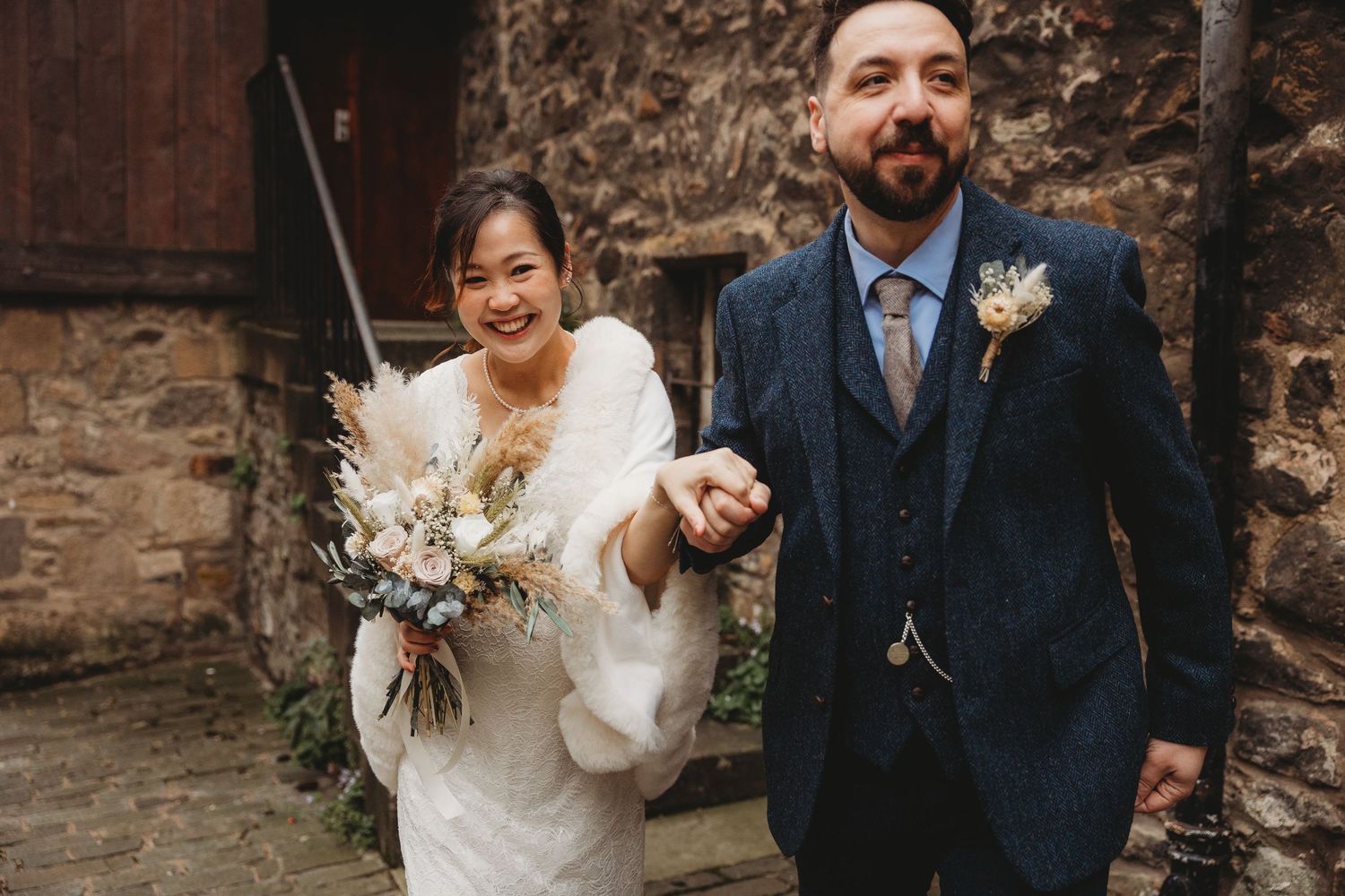 Newlywed couple smiling and walking together outdoors in wedding attire with bride holding bouquet against stone wall backdrop.