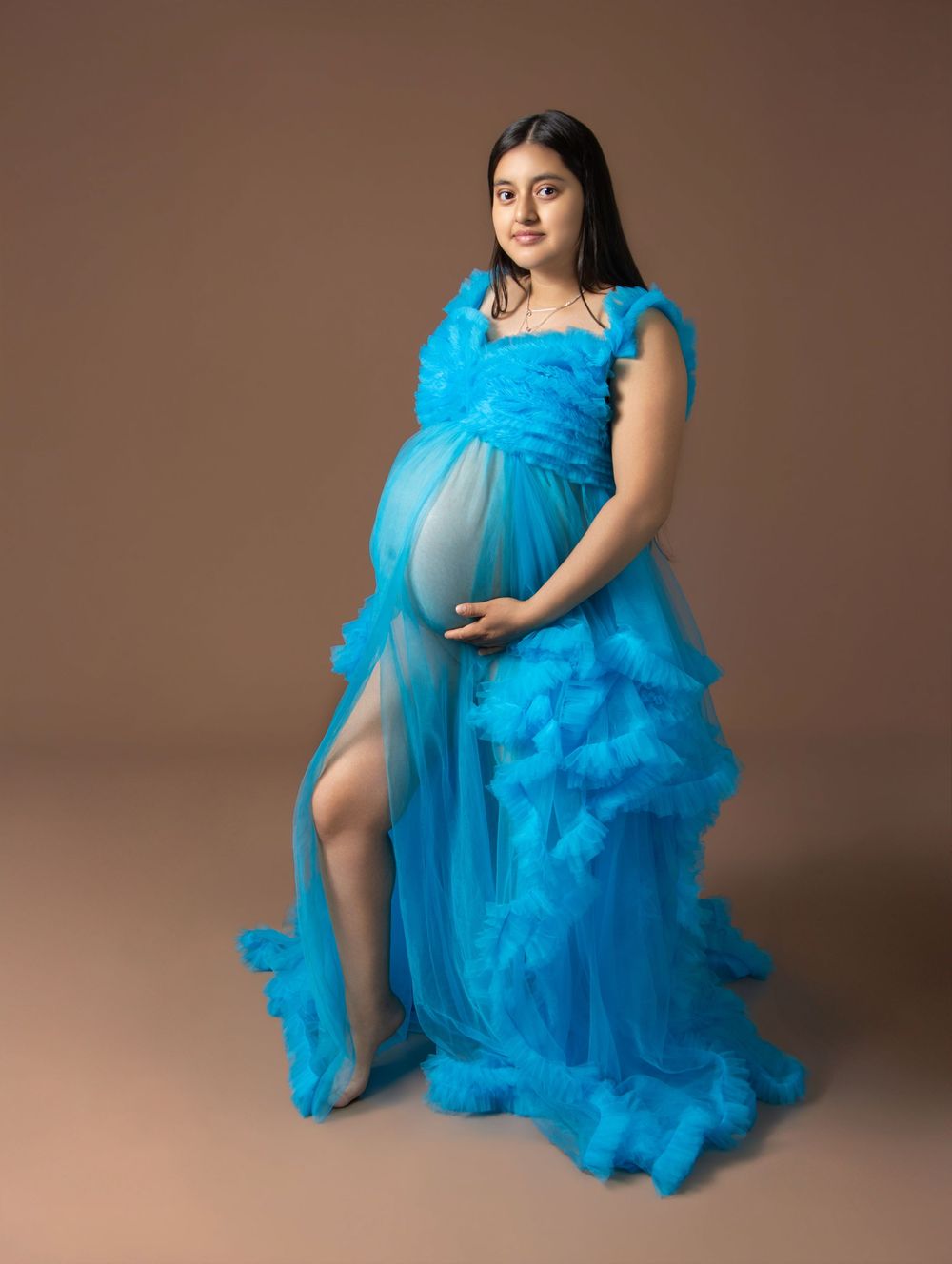 Pregnant woman in a stunning blue ruffled tulle maternity gown posing gracefully in a professional studio setting.