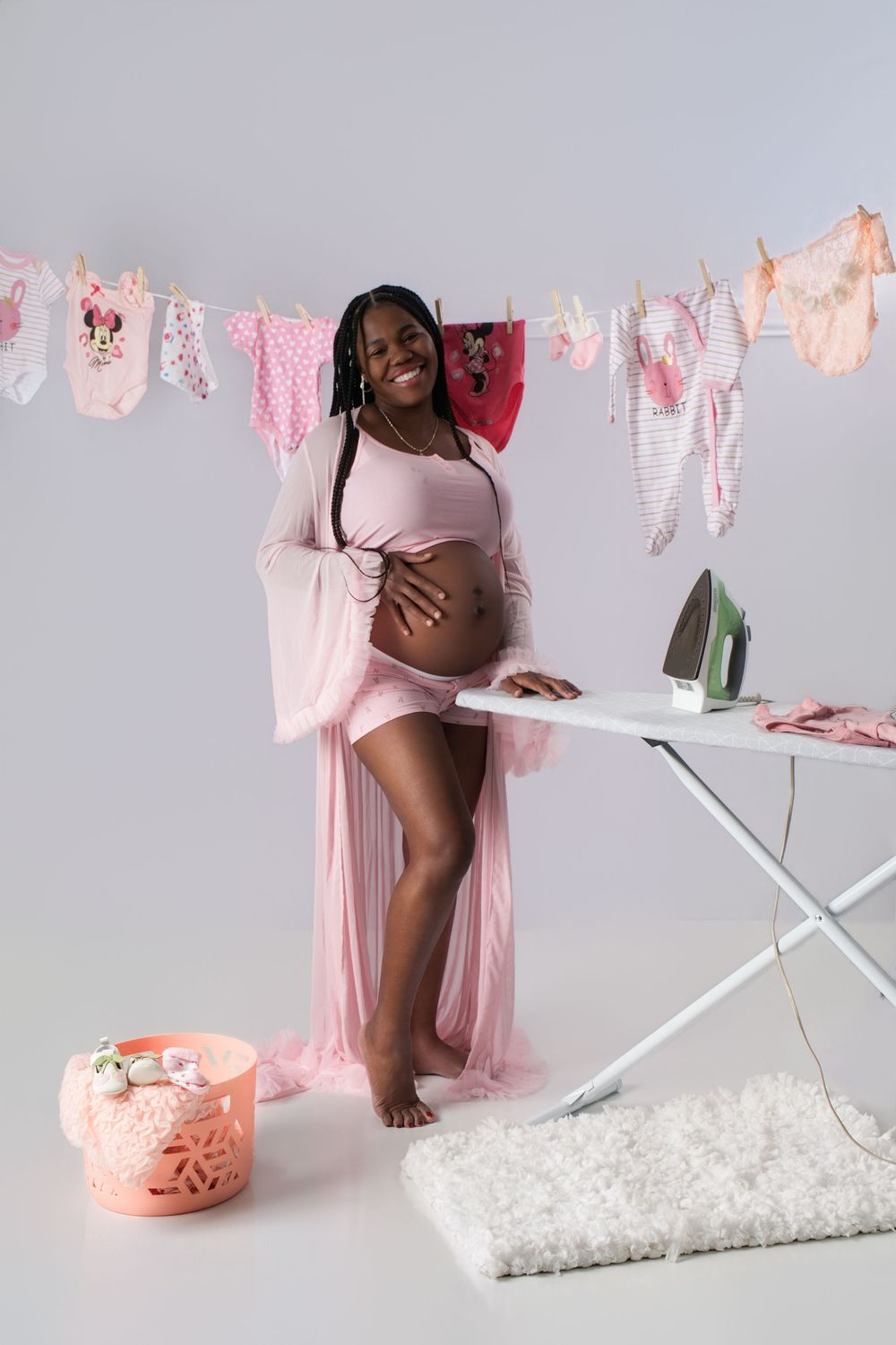 Pregnant woman in pink robe stands by ironing board with baby clothes on a clothesline in a studio setting.