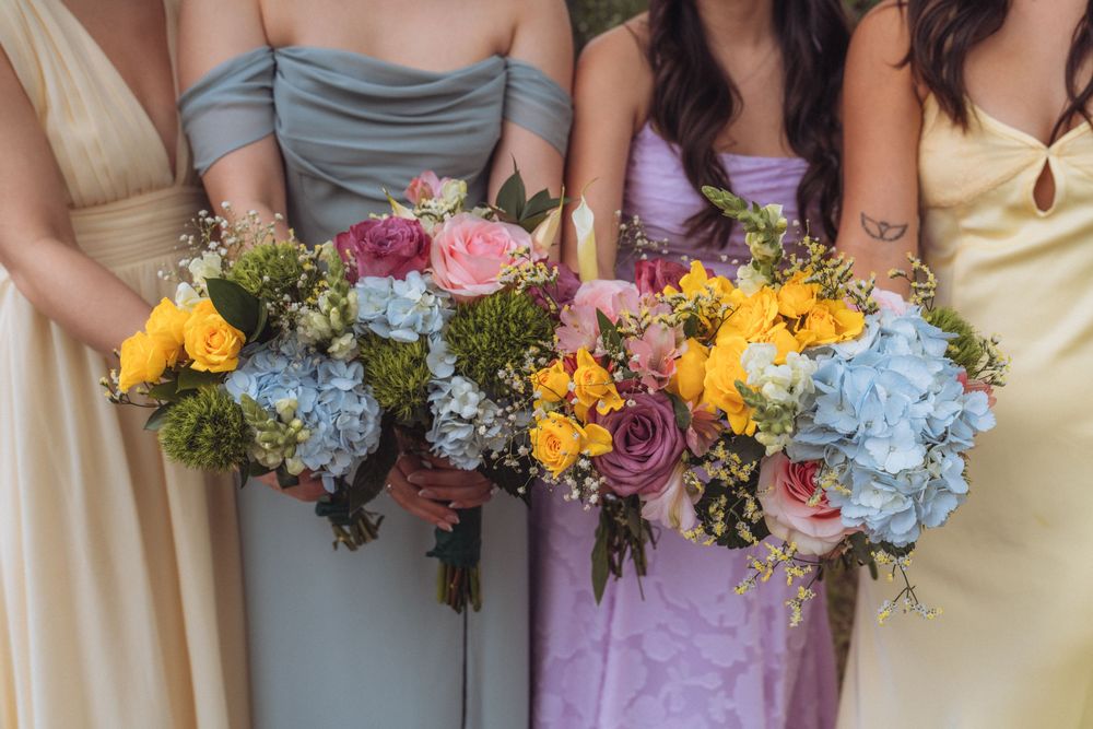 Bridesmaids in pastel gowns hold colorful wedding bouquets featuring blue hydrangeas, pink roses, and yellow flowers.