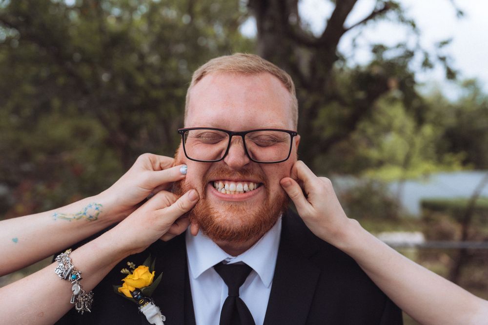 Redheaded groom in black suit smiles as hands pinch his cheeks at outdoor wedding.