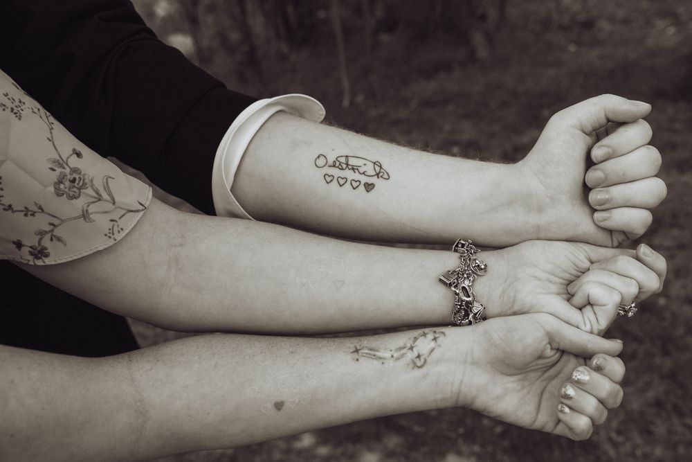 Close-up of three arms with tattoos and bracelets displayed side by side in a black and white photo.