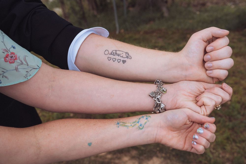 Close-up of two arms showing matching tattoos, a bracelet, and rings at an outdoor wedding.
