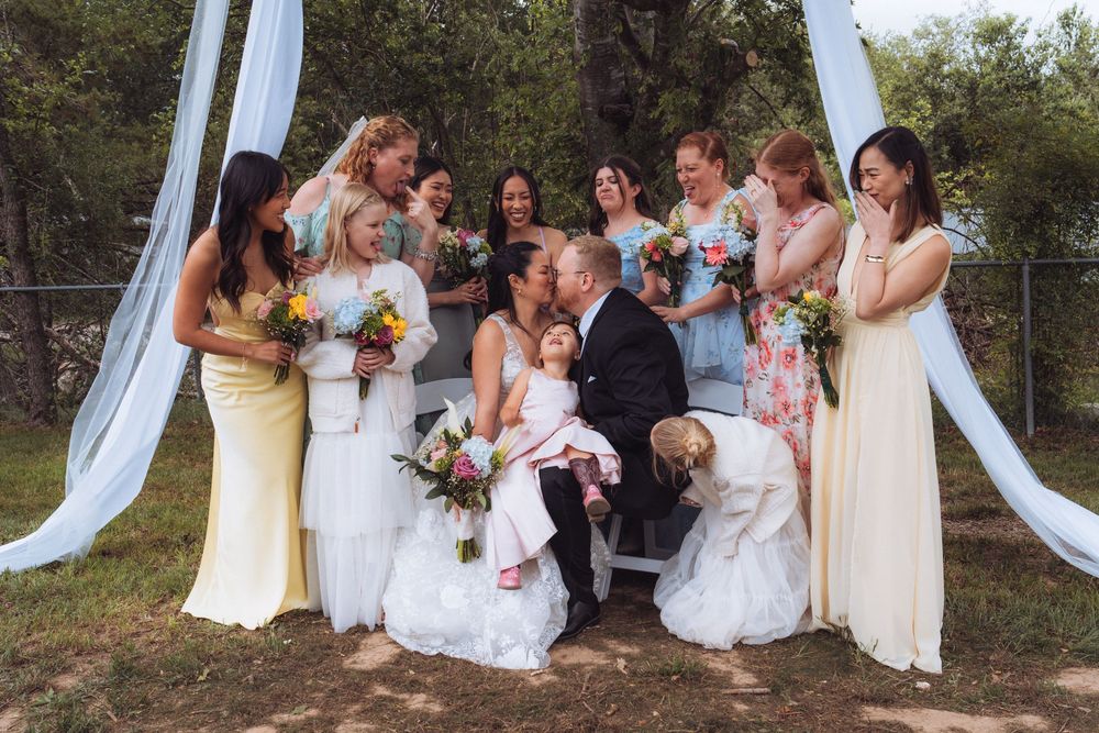 Wedding party with bride and groom sharing a kiss surrounded by bridesmaids in yellow and floral dresses holding colorful bouquets.