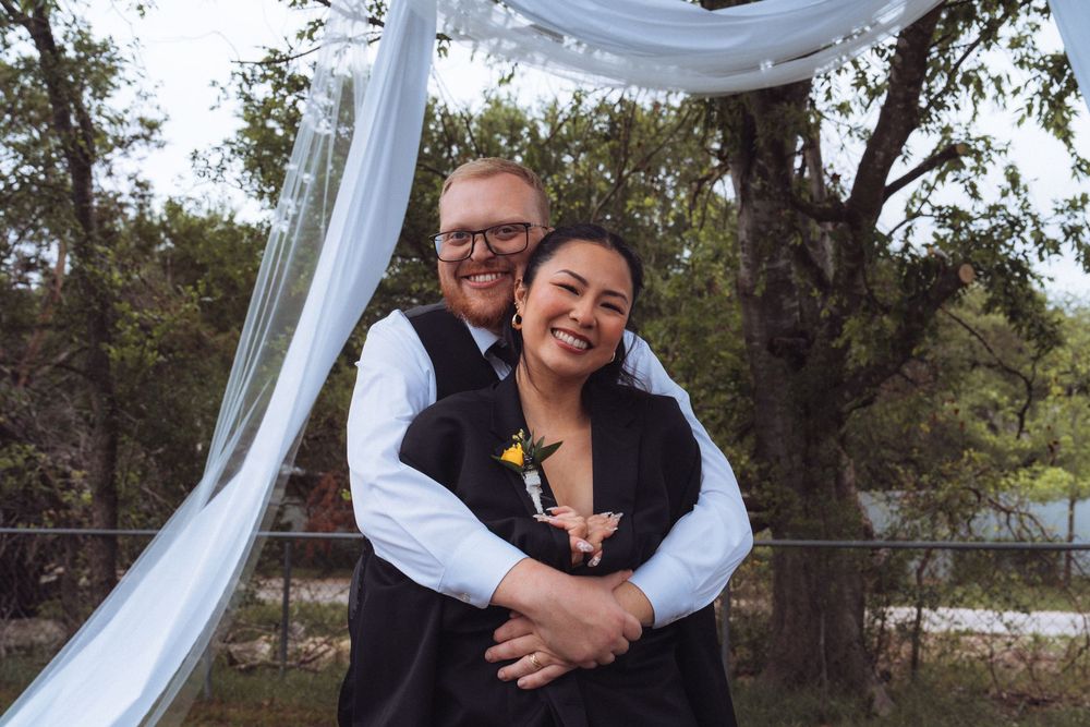 Happy couple embraces outdoors under white draped arch at rustic wedding ceremony surrounded by trees.