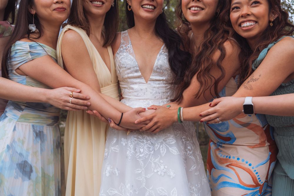 Bride in white lace gown smiling with four bridesmaids in colorful dresses, all holding hands together.