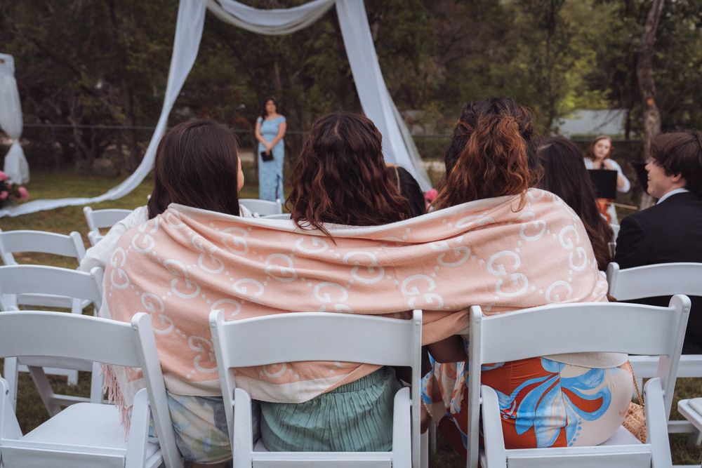 Three women sit together at an outdoor wedding ceremony, sharing a pink blanket on white chairs.