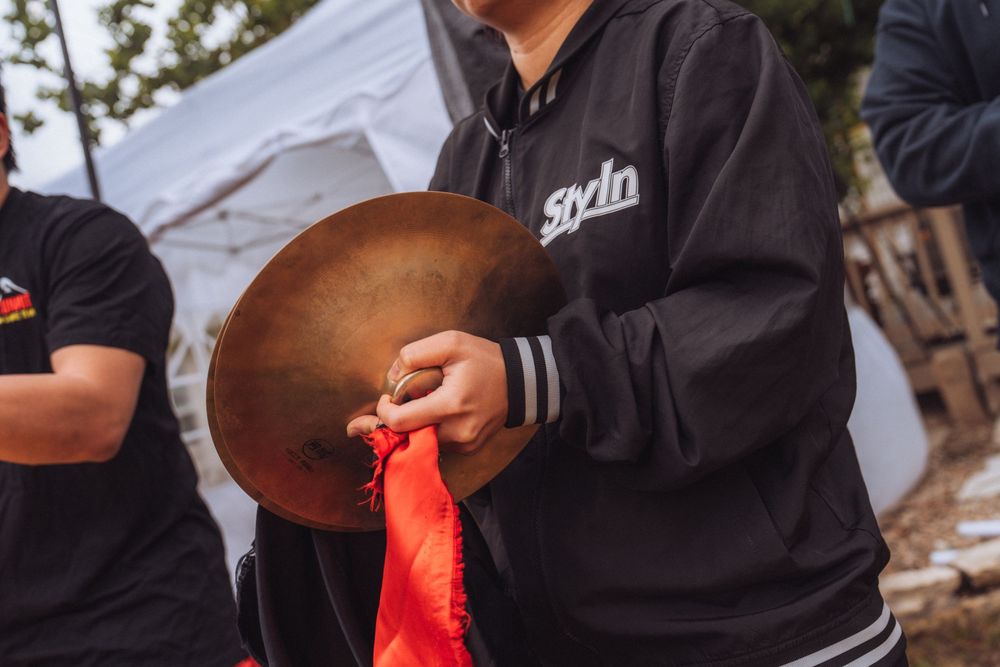 Person in black Stylin hoodie holding a bronze cymbal with red ribbon outdoors near white tent.