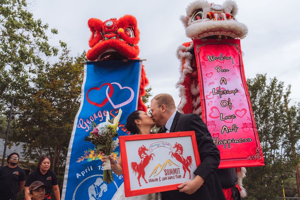 Couple kissing at Chinese lion dance wedding celebration with colorful banners and traditional dragon decorations outdoors.