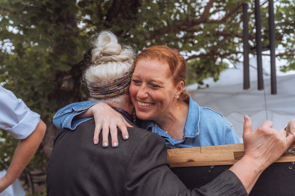 Two women sharing a warm hug outdoors, one with red hair smiling broadly, surrounded by trees.