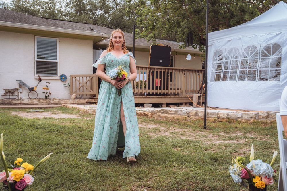 A woman in a teal floral gown holds a bouquet at a backyard wedding ceremony.