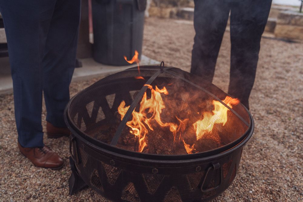 A metal fire pit with vibrant orange flames burning outdoors, with two people standing nearby on a gravel surface.