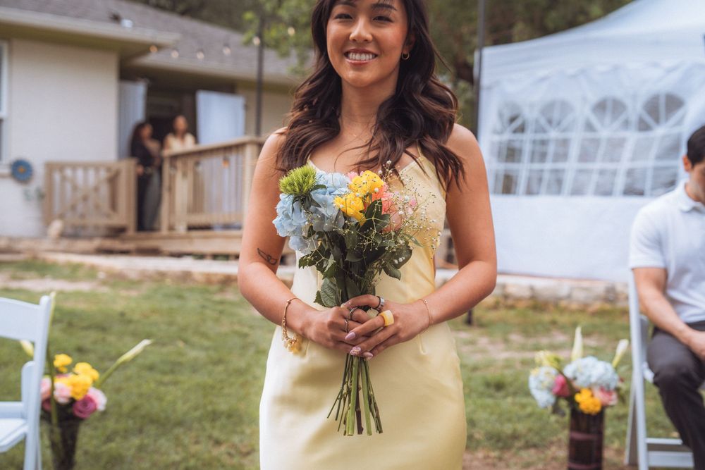 Smiling bridesmaid in yellow dress holds colorful bouquet at outdoor wedding ceremony.