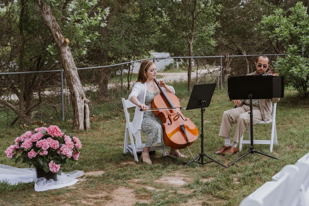 Two musicians play cello and violin outdoors at a wedding ceremony surrounded by lush green trees.