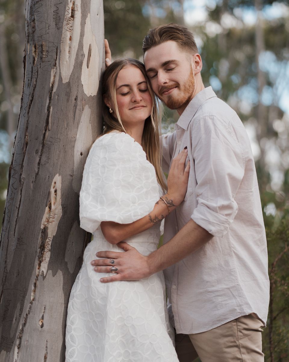 Couple embraces tenderly against a eucalyptus tree, the woman in a white dress and the man in a beige shirt engagement photoshoot rouse hill