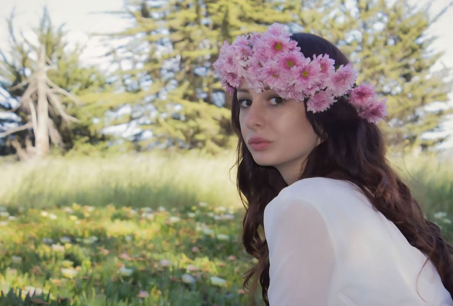 A young woman wearing a crown of flowers poses near a field of flowers.
