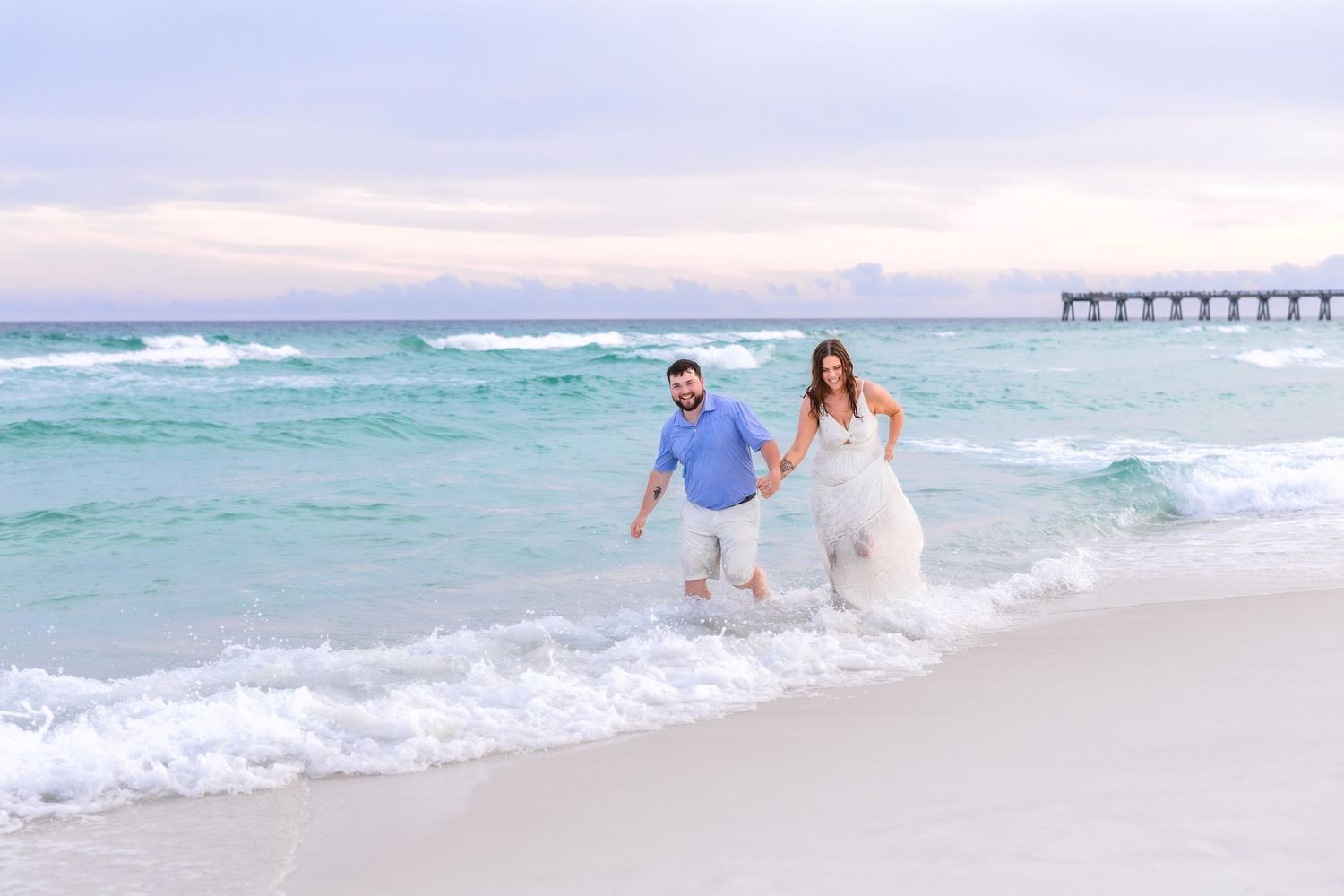 Couple in wedding attire walking hand-in-hand along a turquoise beach shoreline with a pier in the background.
