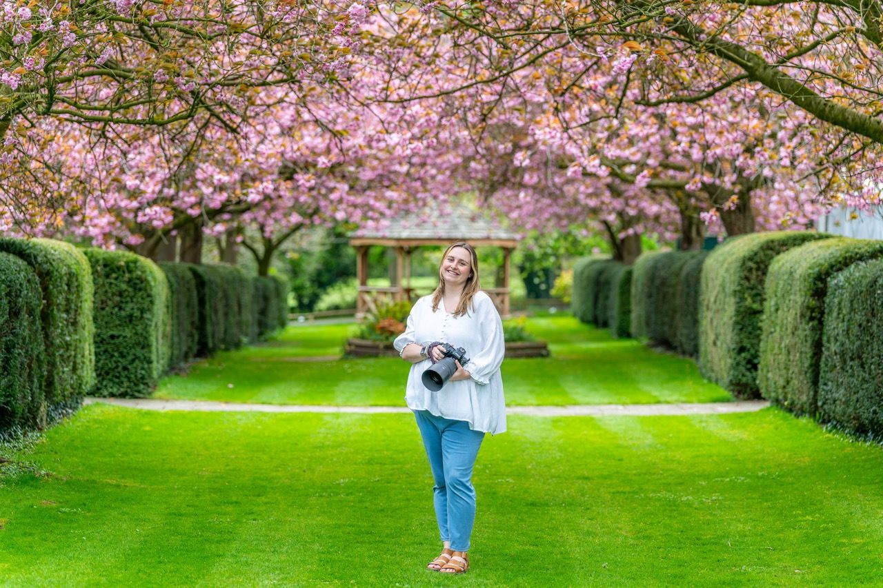 Woman holding camera standing in garden path lined with pink cherry blossom trees and trimmed hedges on green lawn.