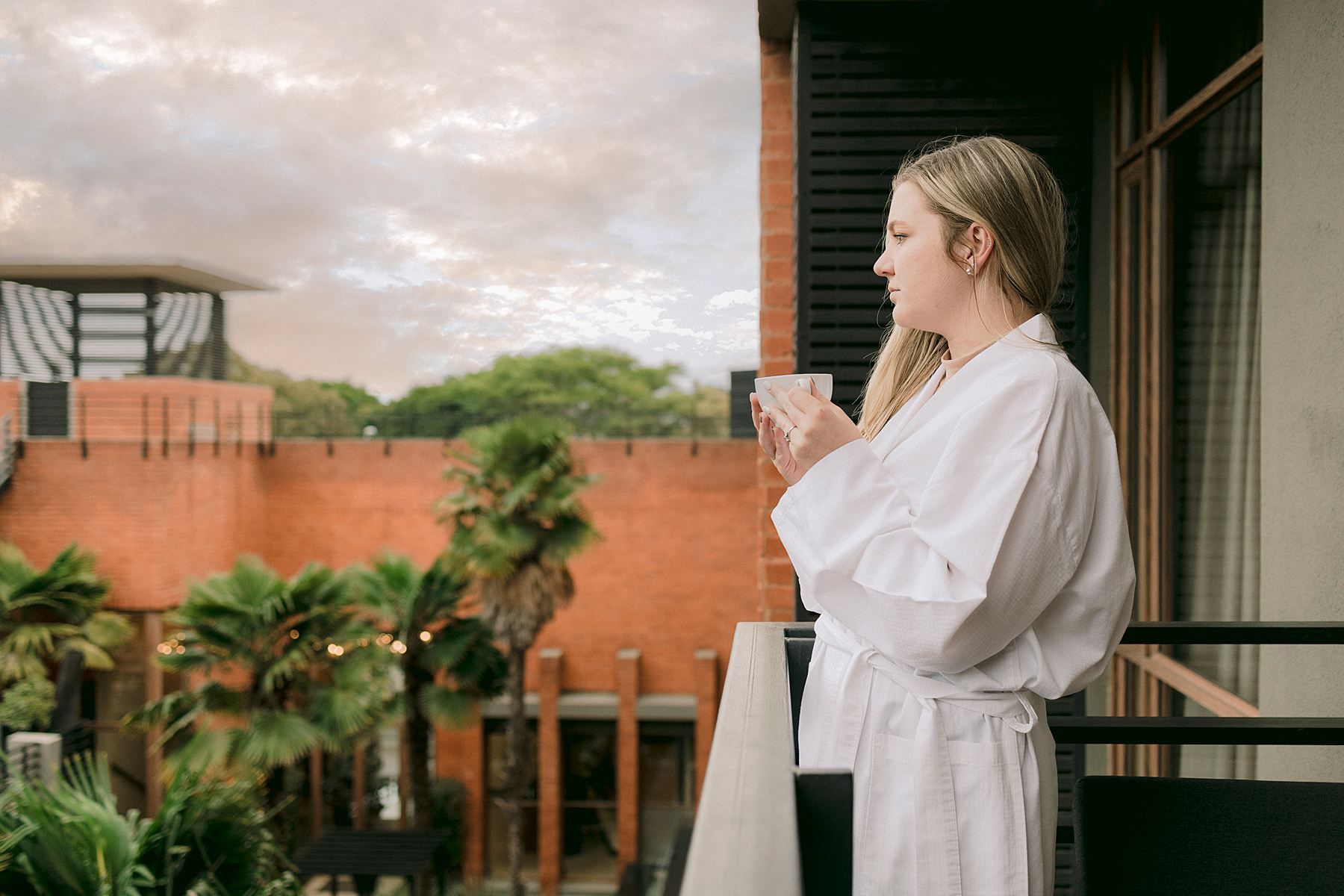 Woman in white bathrobe holding drink on hotel balcony at sunset with tropical garden view.