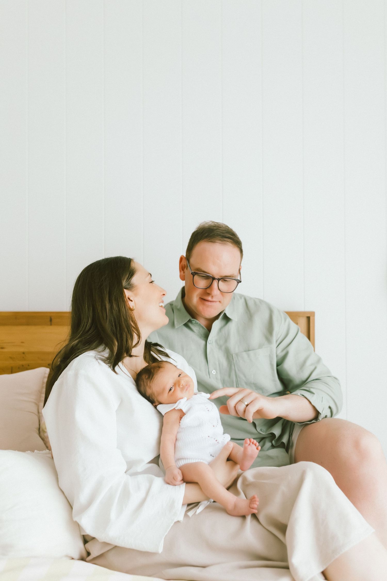 Happy parents sitting on bed holding their newborn baby, smiling at camera in bright airy bedroom.