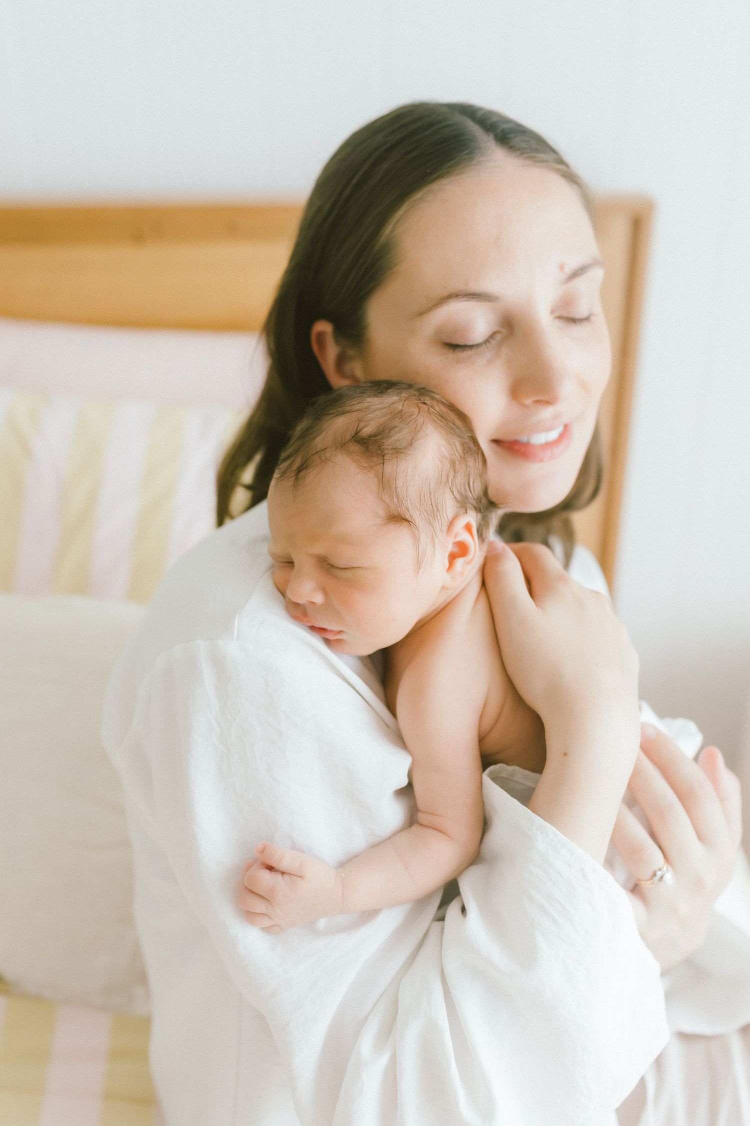 A mother in a white robe tenderly holds her sleeping newborn baby close to her chest in a bright, serene room.