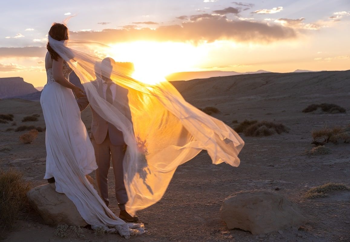 Bride in flowing white gown and veil blowing in wind at desert sunset landscape.