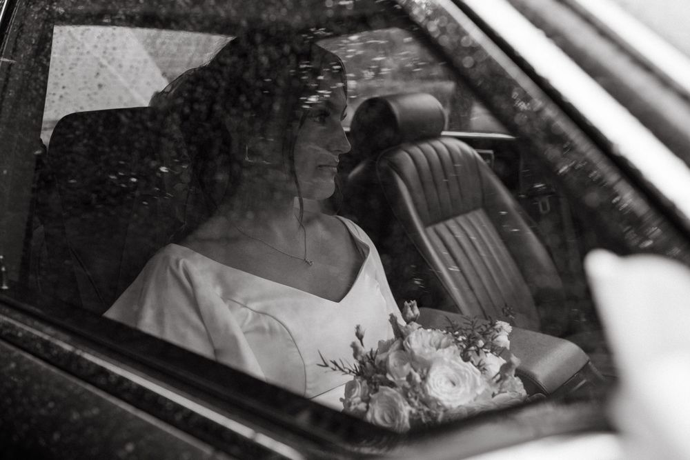 bride sitting in a vintage car looking at raindrops on the window while being captured by storytelling wedding photographer in Uppsala