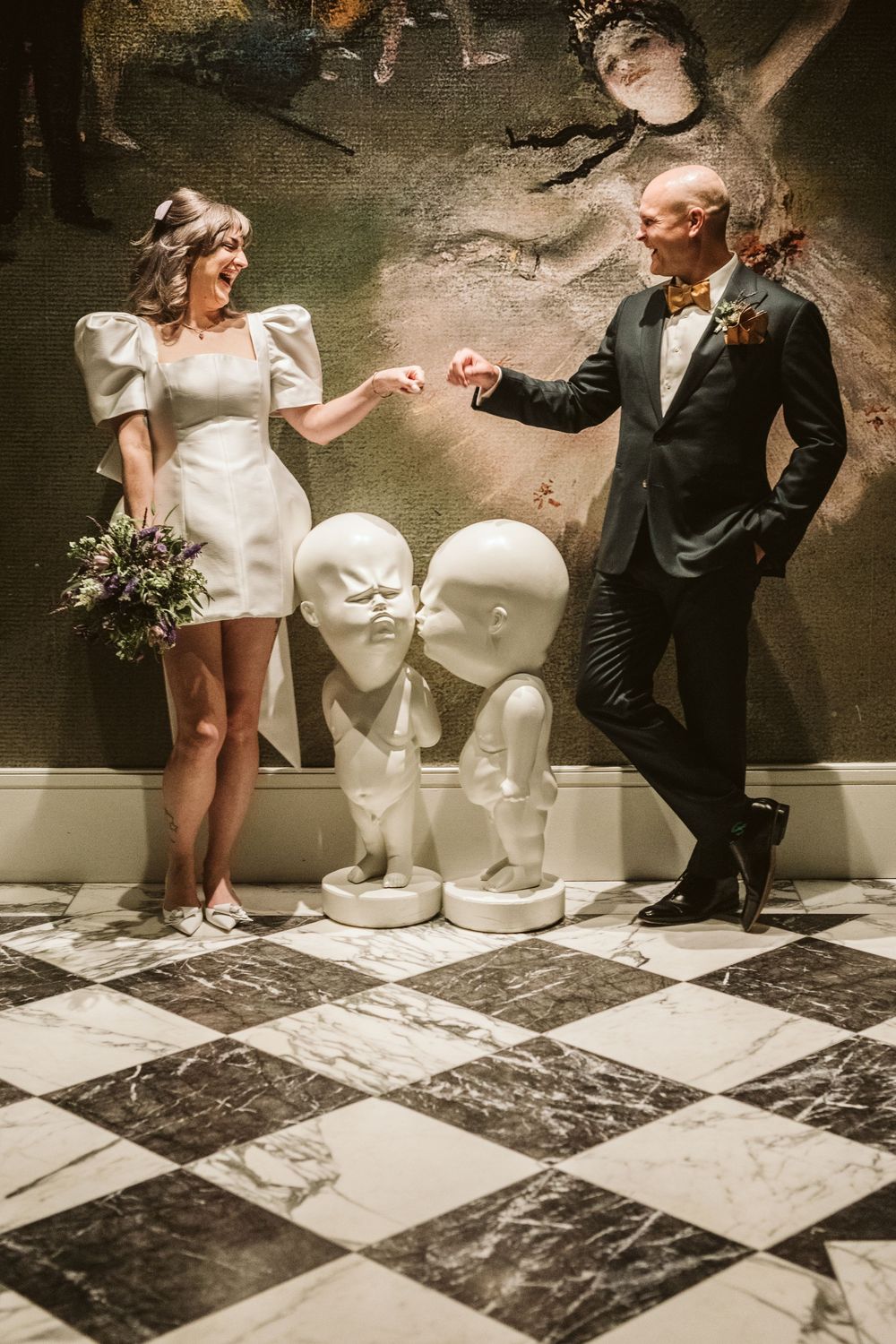 Bride and groom holding hands beside white sculptural busts on a checkered marble floor with a dramatic painted mural backdrop.