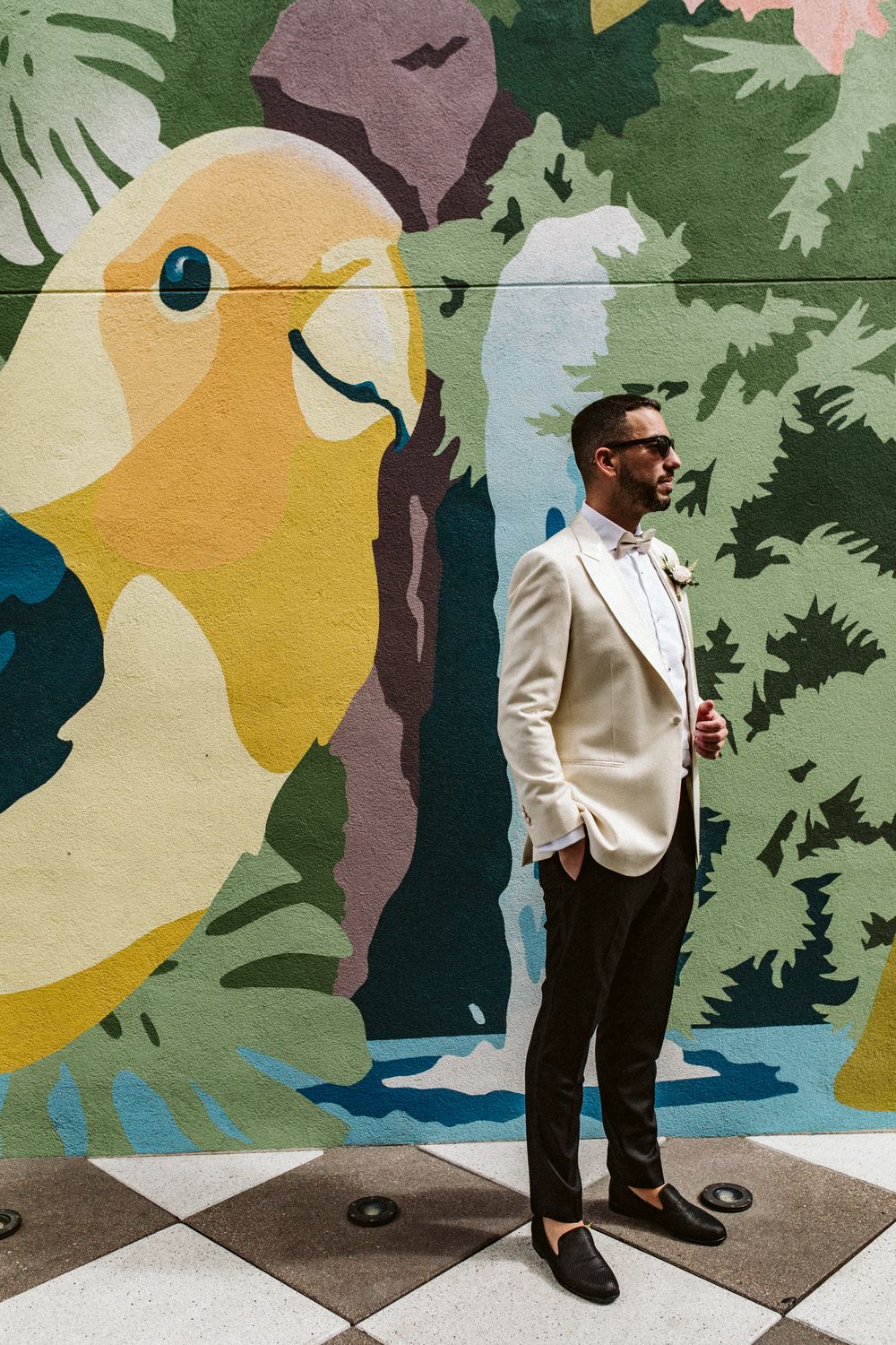Groom in white tuxedo jacket and bow tie poses before a vibrant tropical bird mural on a checkered floor.