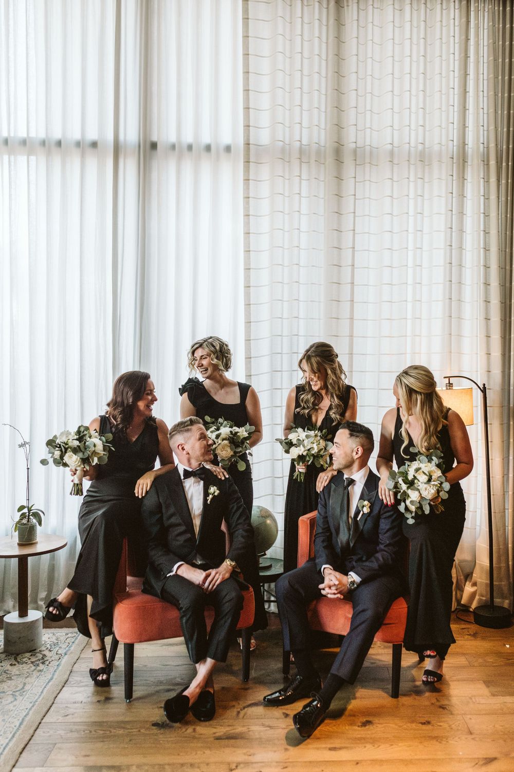 Wedding party in black attire, two grooms seated in red chairs with bridesmaids holding white bouquets near sheer curtains.
