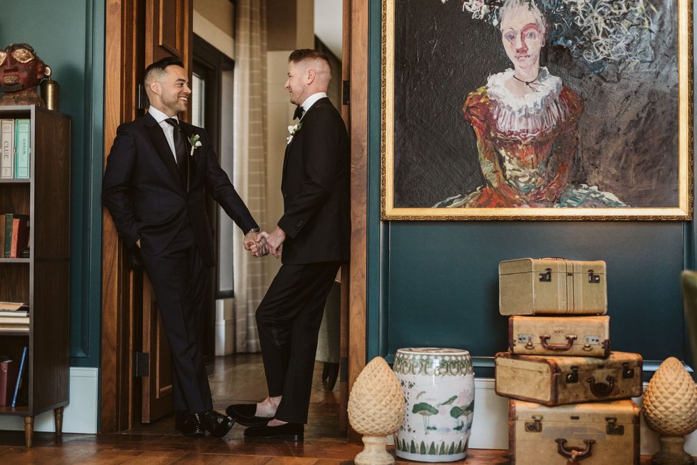 Two grooms in black tuxedos hold hands in an elegant room with vintage trunks and a classic portrait painting.