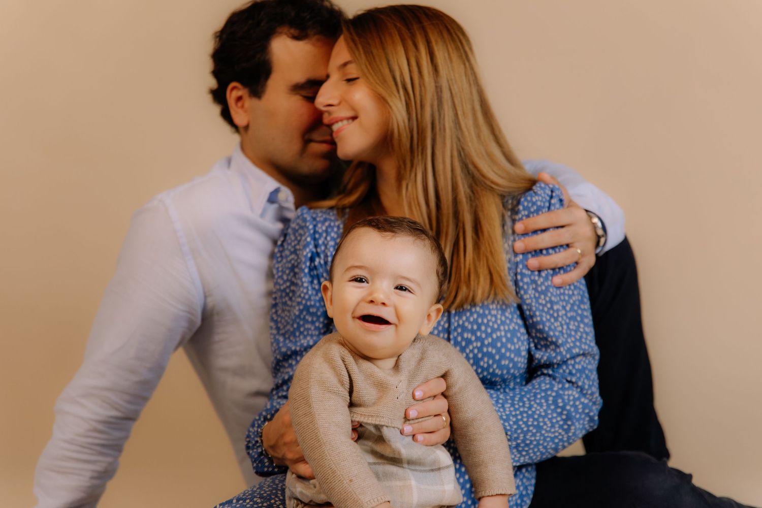 Happy family portrait with parents smiling and kissing while holding their laughing baby in a studio setting.