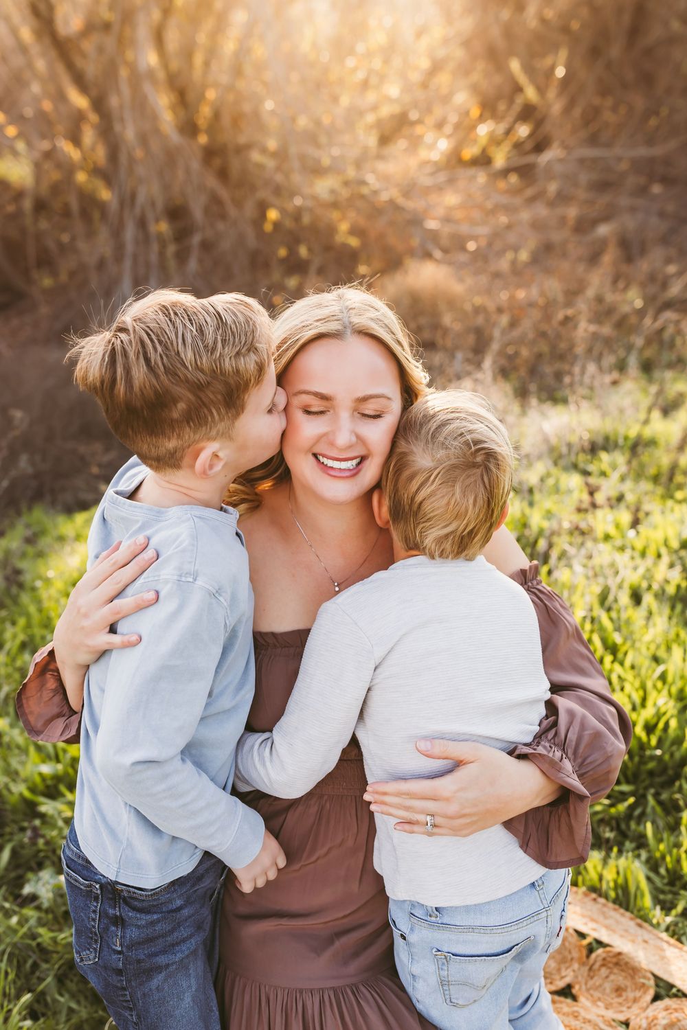 A smiling mother hugs her two young sons outdoors in golden sunlight, one boy kissing her cheek.
