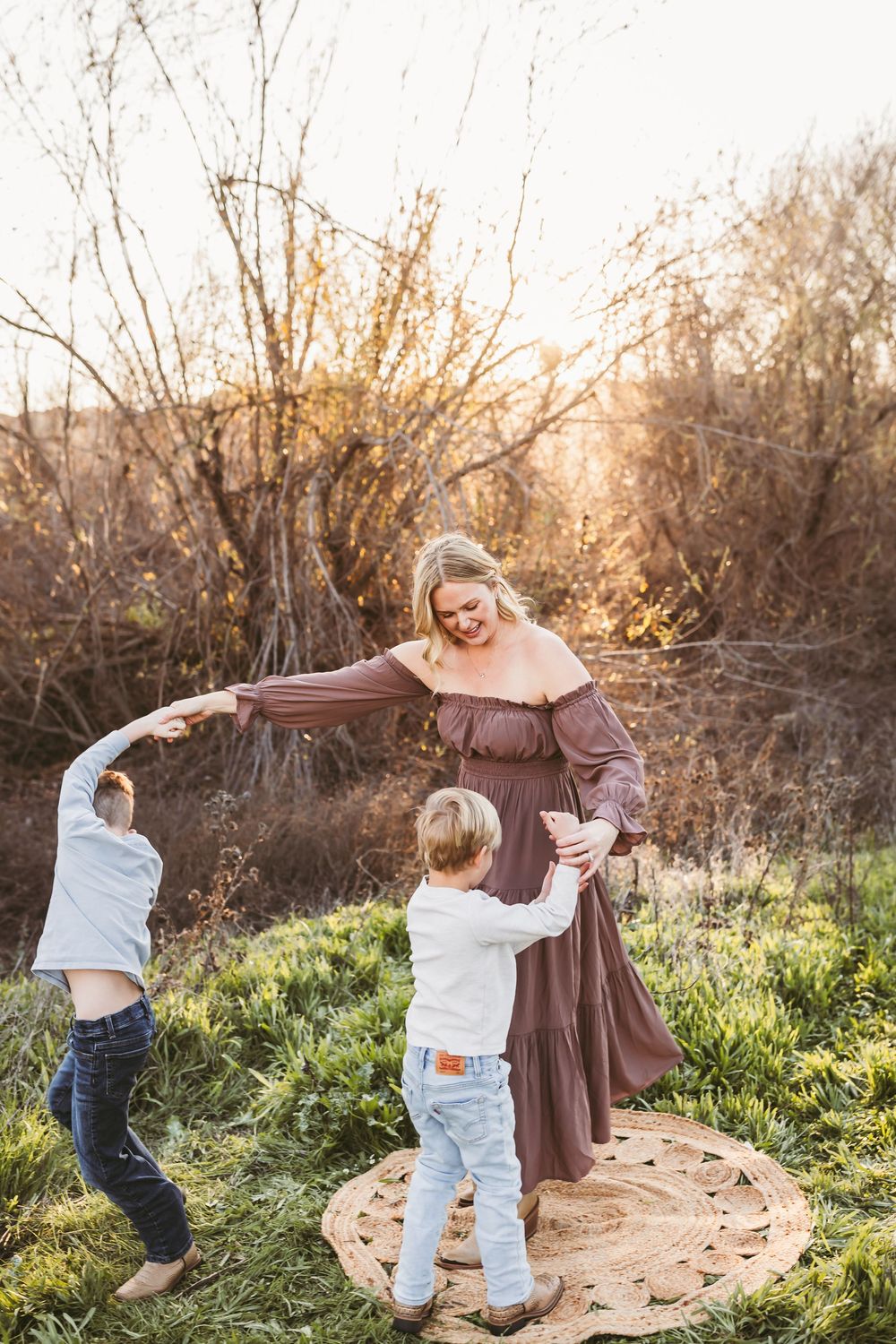 A woman in a mauve dress dances outdoors with two young children at golden hour.