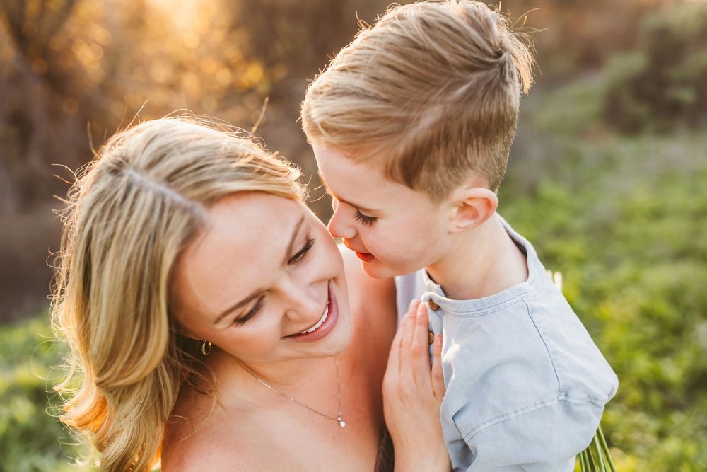 A joyful mother laughs as her young blond son whispers in her ear outdoors in warm golden sunlight.